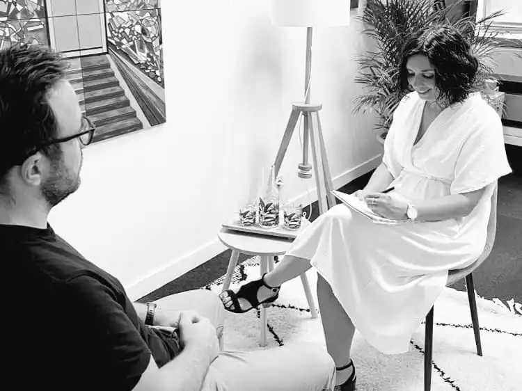 Lola MONACO et son client, dans son bureau, assis sur des fauteuils. Lola MONACO sourit et prend des notes